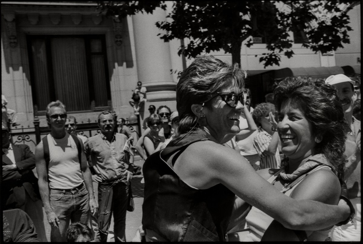 Two woman smile as they greet one another in a black and white photo, one woman has a leather vest and sunglasses and teased, short blonde hair. The other has fanned out brunette hair.