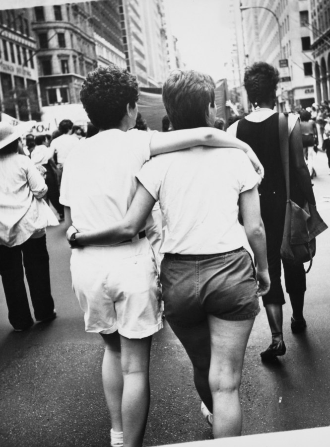 A photo of two women walking while having their arms wrapped around each other, shot from behind in black and white.