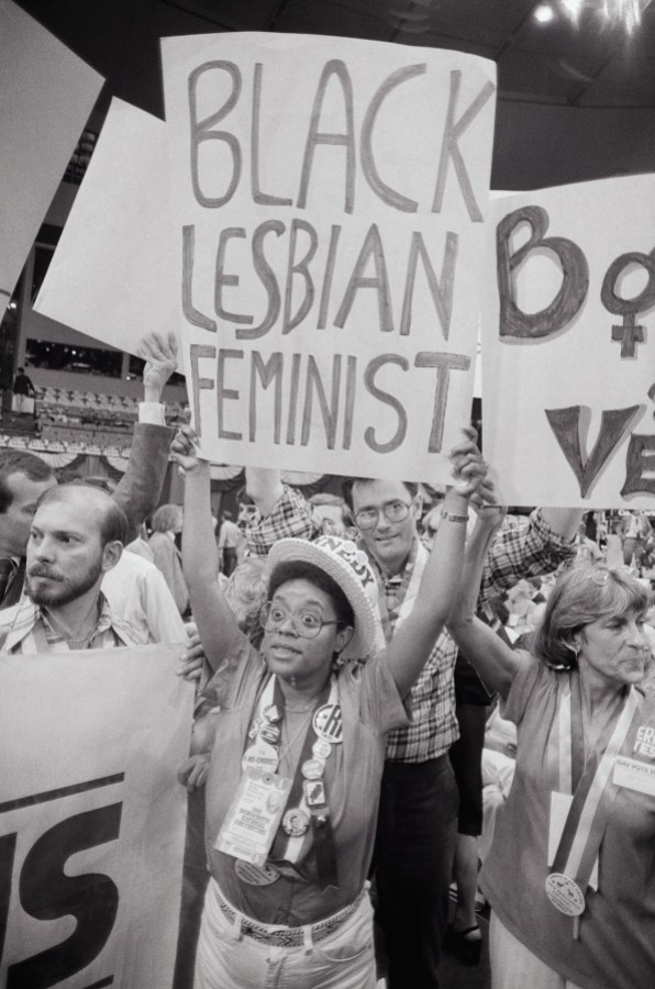 In a black and white photo, a black woman wears a hat and has on a lot of conference buttons around a lanyard. She holds up a sign that says "Black Lesbian Feminist"