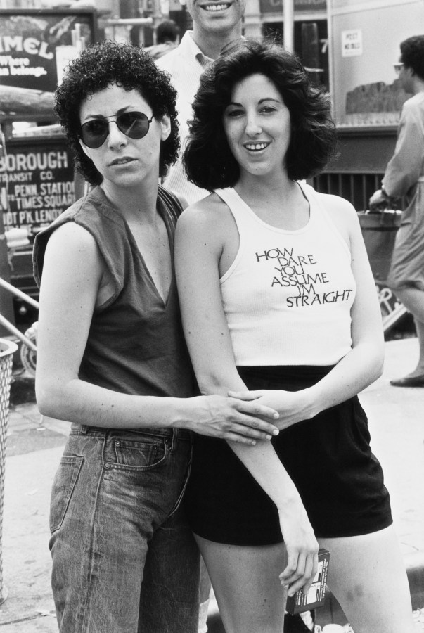 In a black and white photo, a white lesbian couple embraces each other during Pride. One has on a shirt that says How dare you assume I'm straight'. 