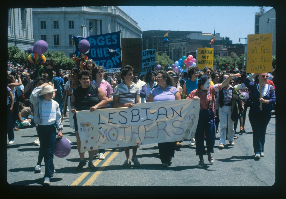 In a color photo with a black edge around all sides, A group of women march behind a banner reading "Lesbian Mothers" during the Lesbian/Gay Freedom Parade in San Francisco, CA.