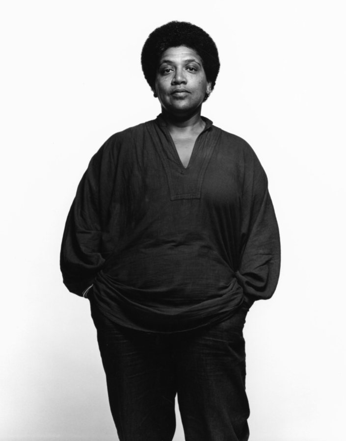 A black and white photo of Audre Lorde with a short afro and a dark colored v-neck collard shirt with her hands tucked into her pockets. She looks serious. She is against a white background.