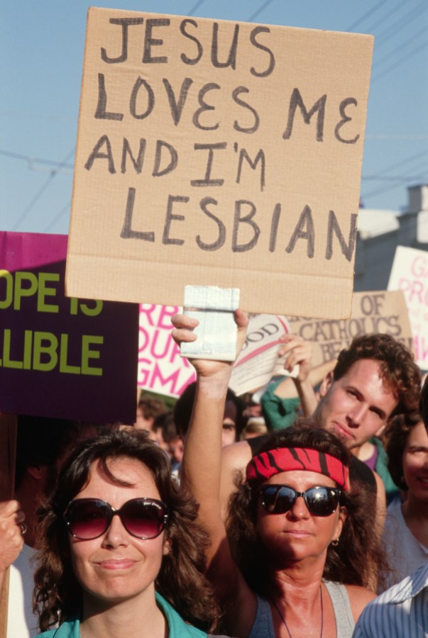 A woman with sunglasses and a red bandana holds a sign that says "Jesus Loves Me and I'm a Lesbian"