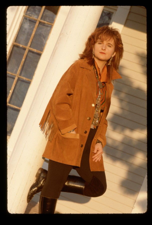 Melissa Etheridge in a brown camel coat leaning against a porch railing.