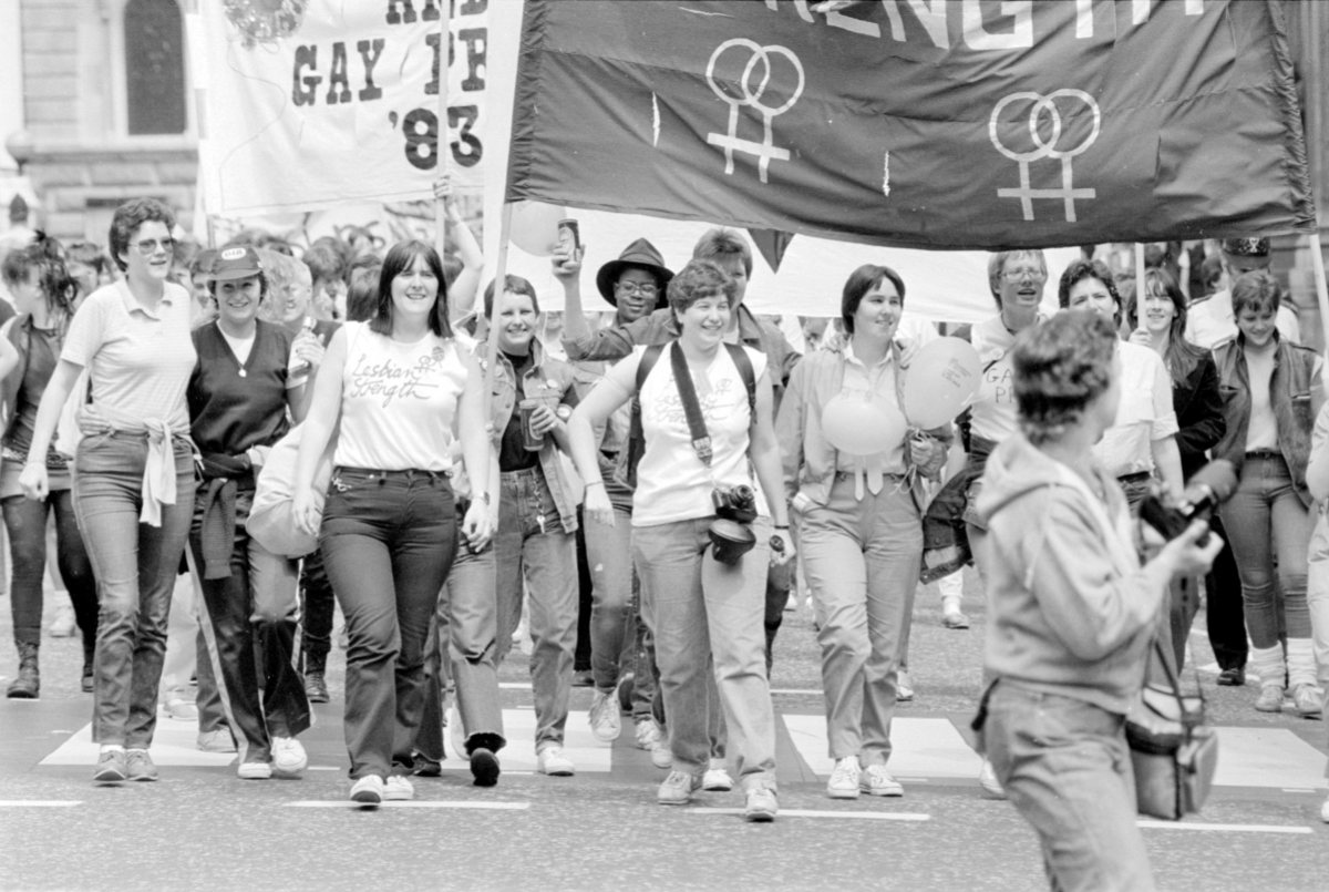 A wide shot, black and white photo, of a large group of lesbians marching during Pride in London, they have large signs that are being cut off by the upper edge of the photo.