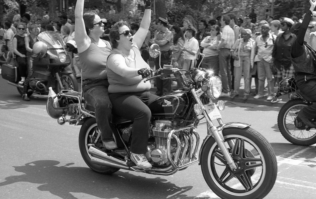 Two fat white lesbians share a motorcycle during the Dyke March in a black and white photo, they have their hands up waving to the crowd.