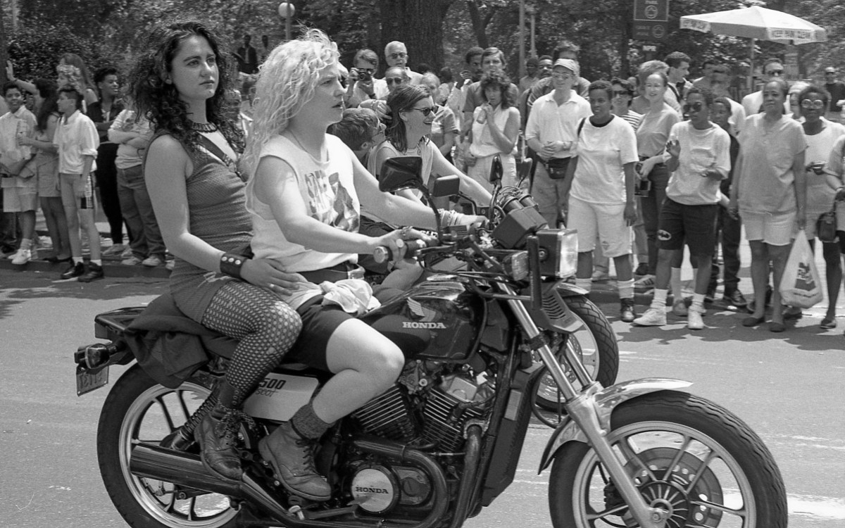 A femme lesbian couple shares a motorcycle. One has permed blonde hair and one has teased brunette hair.