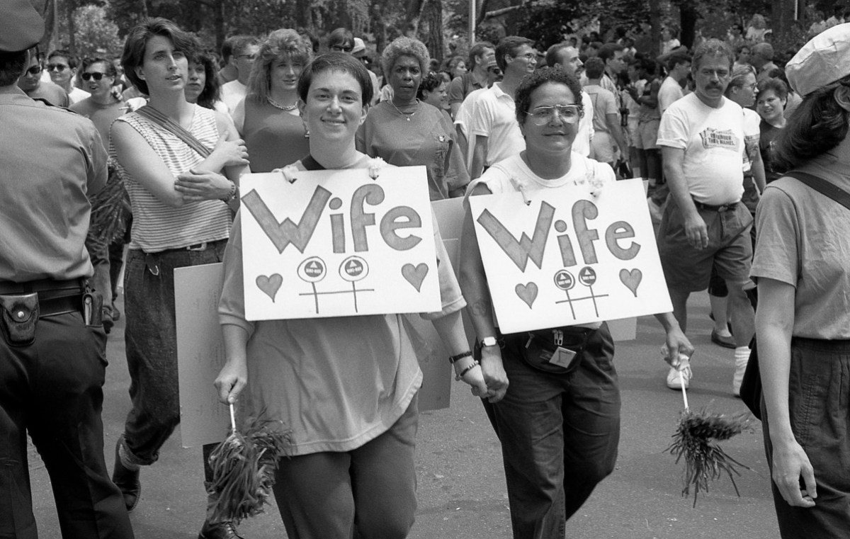 A black and white photo of two lesbians holding hands and marching in a 1989 Pride parade, they both have short brunette hair. They have signs across their chests that say "wife" with hearts underneath.