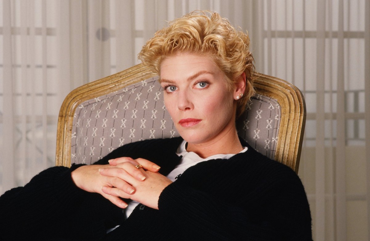 A full color portrait of Kelly McKillis, a white actress with short blonde hair, sitting in a wicker chair with an oversized black sweater.