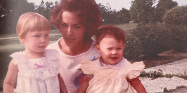 An old photograph of a young, toddler Heather, her baby sister, and their mom holding them against a landscaped background. The young girls are wearing frilly dresses, and the mother's hair is done up in an 80's style. Heather is blonde and he mother and sister are brunette