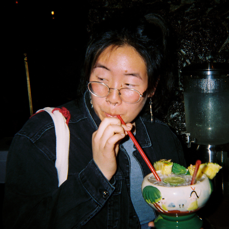 Julie is an East Asian person in glasses and blue shirt underneath a black coat drinking a large fruity drink