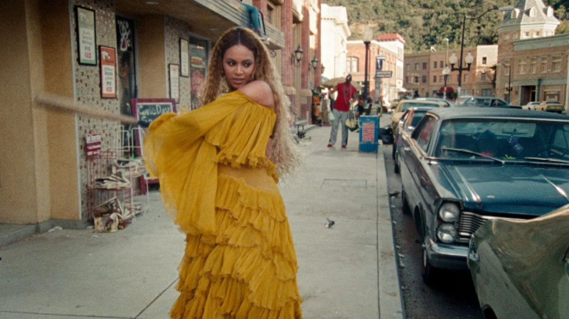 Beyoncé in a long yellow dress swings a baseball bat at a car window