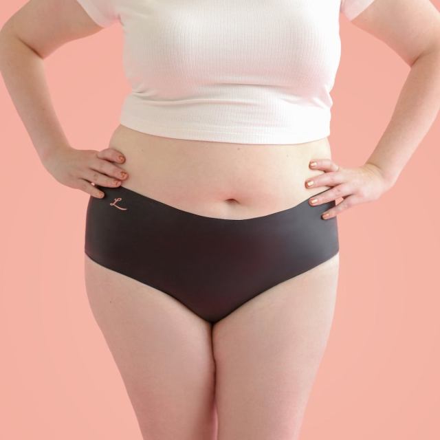 A white person in a white crop top and black latex Lorals underwear stands with their hands on their hips. Only their torso and thighs are visible.