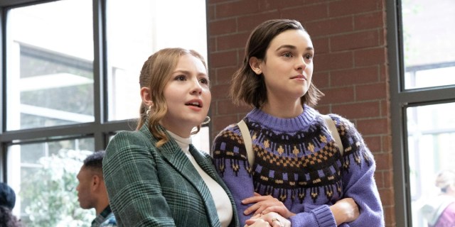 In Love Victor Season 3, Lake and Lucy stand next to each other with their arms crossed in a high school hallway. Lake is in a blue suit coat with a off white turtle neck and hair red hair in a ponytail. Lucy has a blunt brunette bob and a purple knit sweater with a bright pattern.