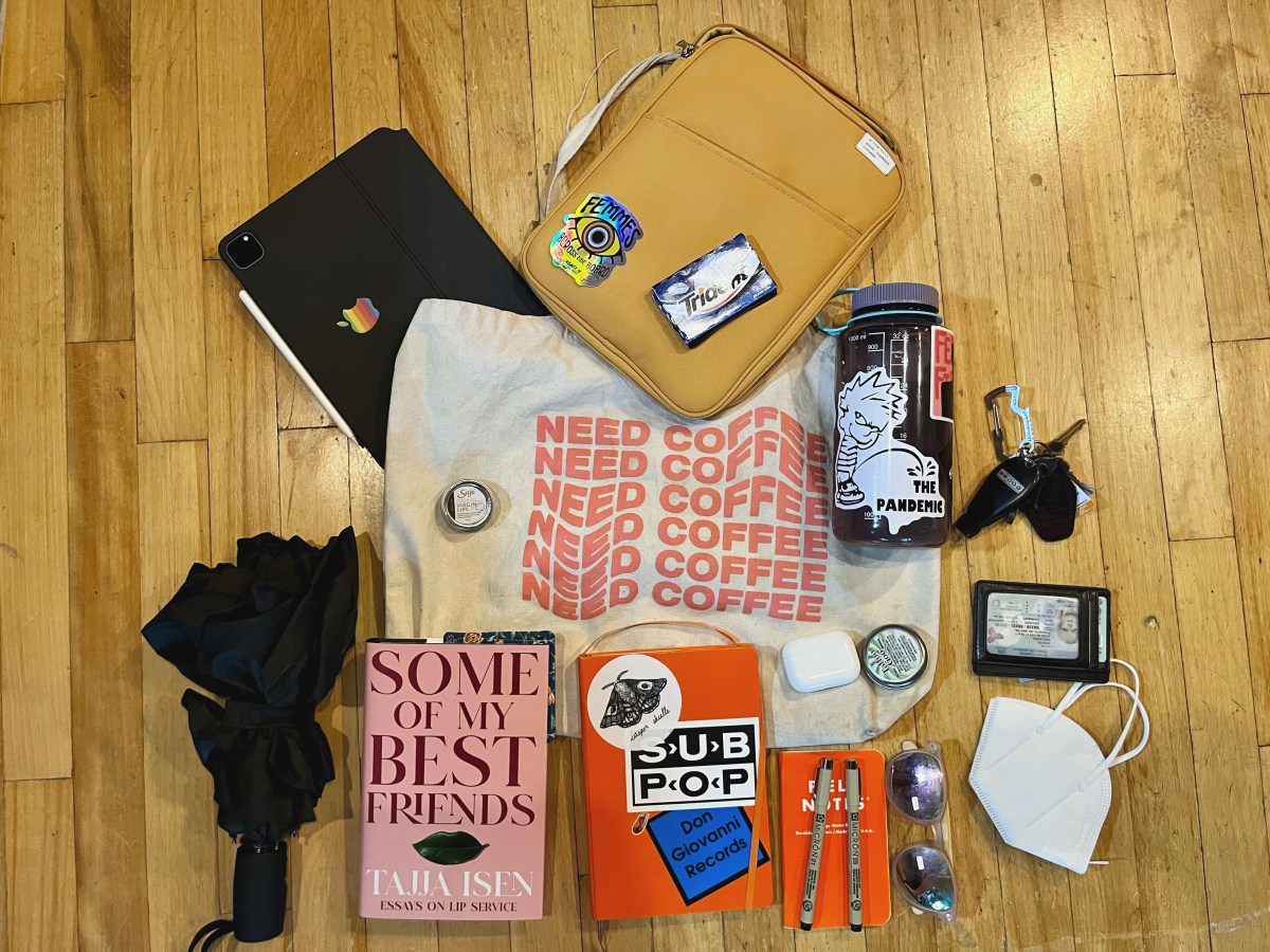 Niko's things are arranged artfully on a wooden surface. There is an umbrella, ipad, book, tote bag that says "need coffee"over and over, small beige leather bag, trident gum, notebook, micron pens, sunglasses, k-95 mask, airpod case, balm, wallet, nalgene and keys.