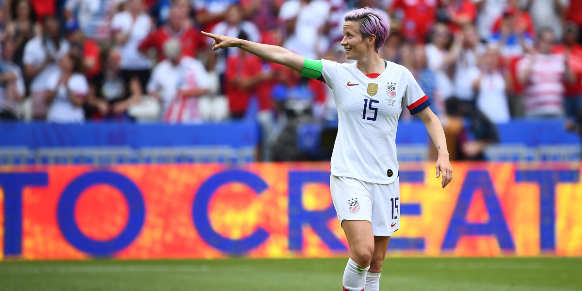 Megan Rapinoe celebrates after scoring in soccer by pointing to the stands. She has on her USWNT uniform and pink hair.