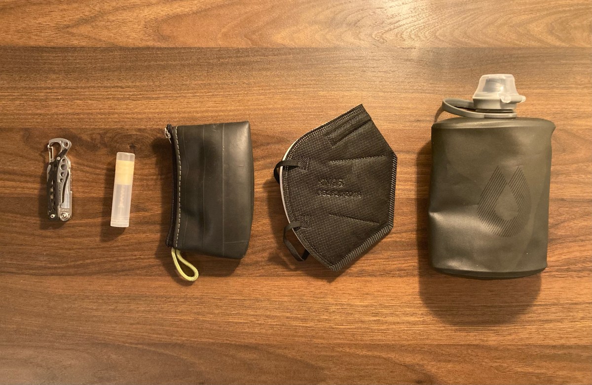 The arrangement of Ro's things. There is a small multi-tool, a homemade chapstic, small weather-proof black bag, black K-95 mask and collapsible water flask, all lined up.