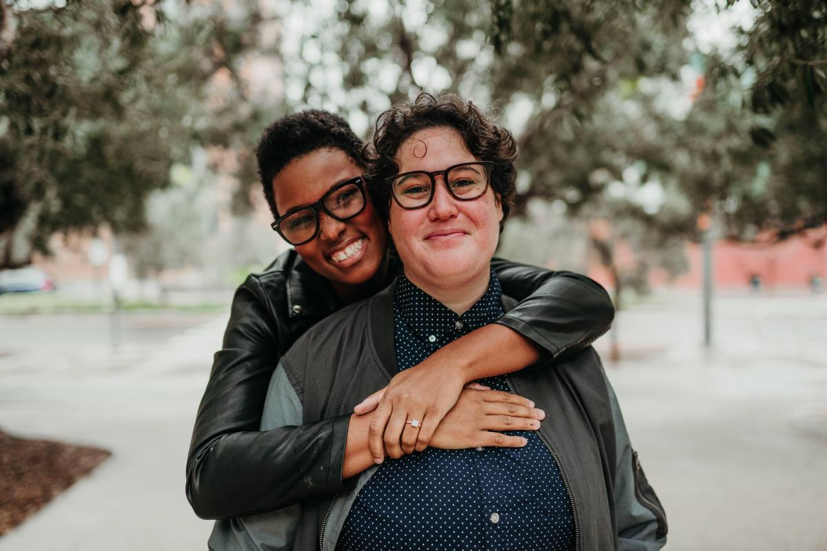 A photo of Sa'iyda and her fiancee, Beth. Sa'iyda stands behind Beth, her arms wrapped around her, revealing an engagement ring on her hand. They both face the camera and smile. Sa'iyda is a Black woman with short black hair wearing glasses and a leather jacket. Beth is a white woman with short curly brown hair wearing a button up shirt, a denim jacket, and glasses.