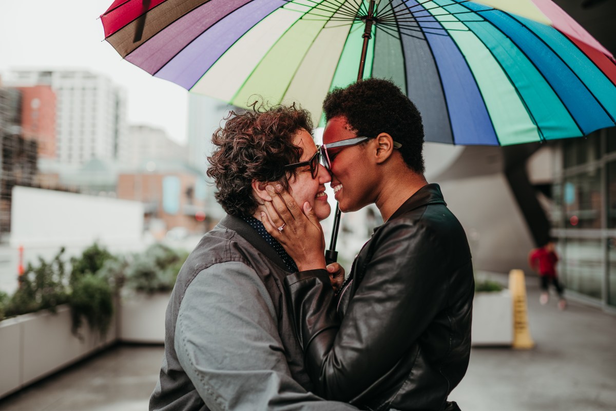 Sa'iyda clasps her fiancee's face in her hands and looks at her lovingly. They stand beneath a rainbow umbrella. Sa'iyda is a Black woman who is wearing blue glasses and a leather jacket and has short hair. Beth, her fiancee, is a white woman with curly short brown hair and black glasses who is wearing a gray jean jacket. An engagement ring is conspicuously visibile on Sa'iyda's hand!