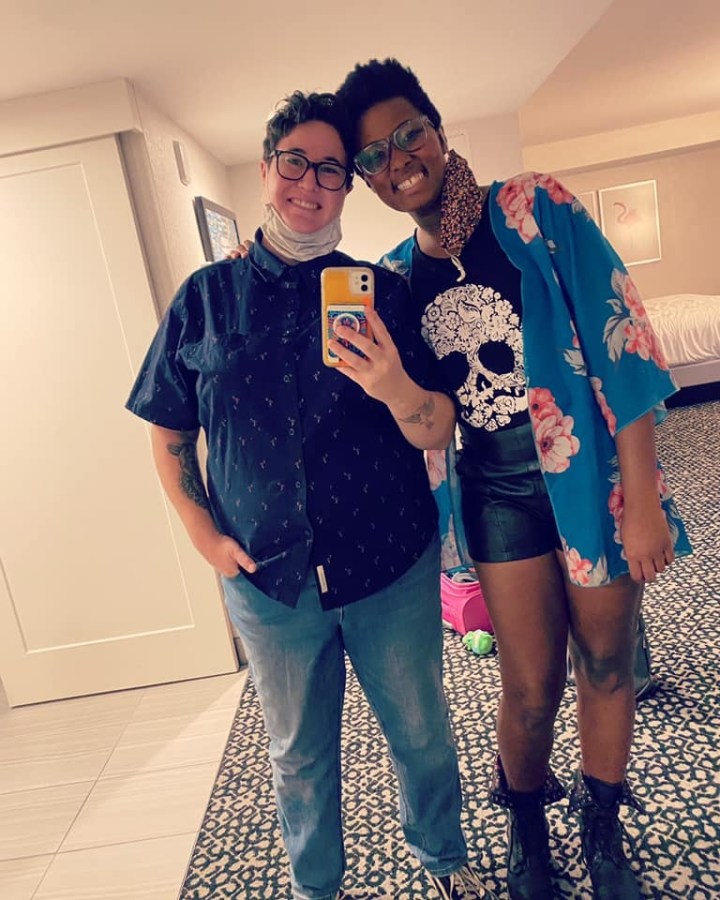 Sa'iyda and Beth bose for a mirror selfie. Beth is dressed in a short sleeved button up and jeans. Sa'iyda is dressed in a skull tee shirt, flowy floral top, what look like leather or pleather shorts, and lace up black leather boots. They both have short hair and glasses and are smiling.