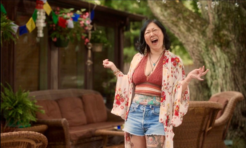 Margaret Cho is in a red knit crop top and a long kimono with red flowers over a beige background, she has on cut off shorts and is standing outside on a deck.