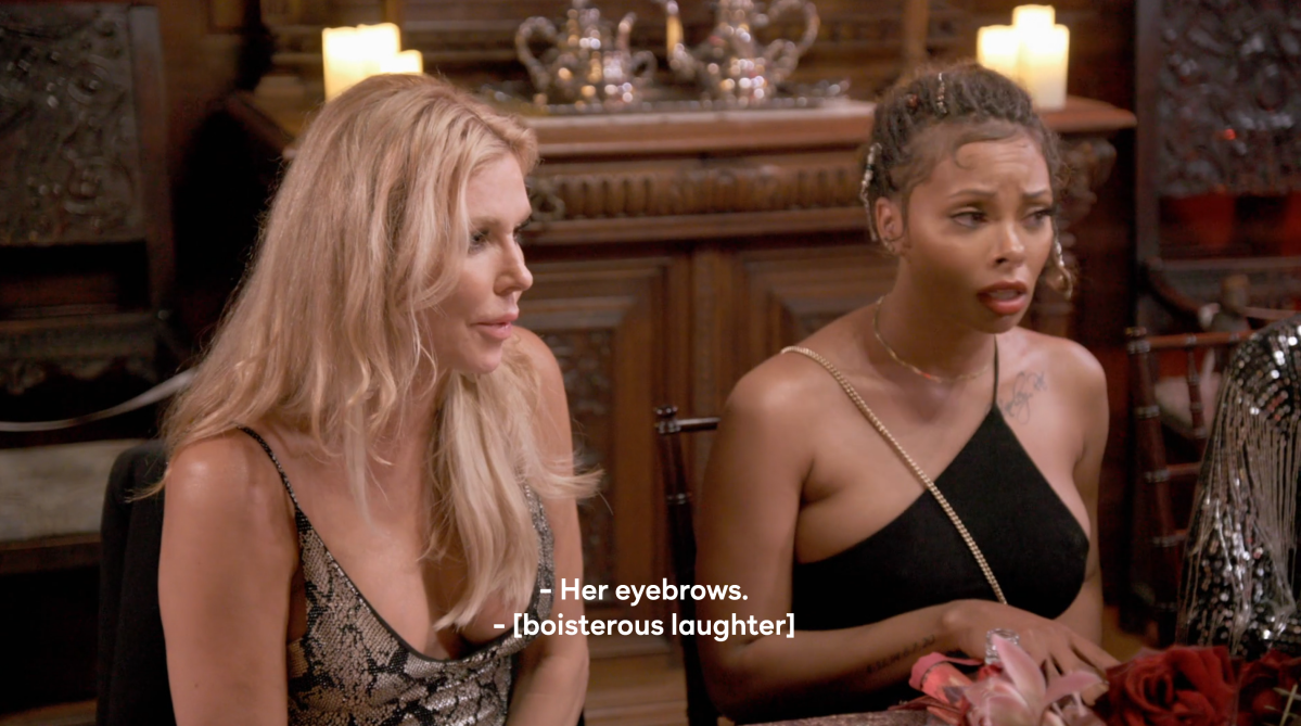 Brandi Glanville, a white woman with blonde hair wearing a snakeskin dress, says "Her eyebrows" on Ultimate Girls Trip. She is sitting next to Eva, a Black woman with a one-shoulder black dress, who looks perplexed.