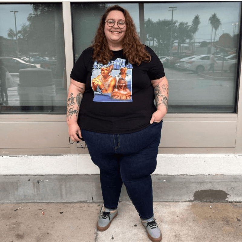 Stef is a white person in black jeans and a black t-shirt with a graphic on it, standing outside.