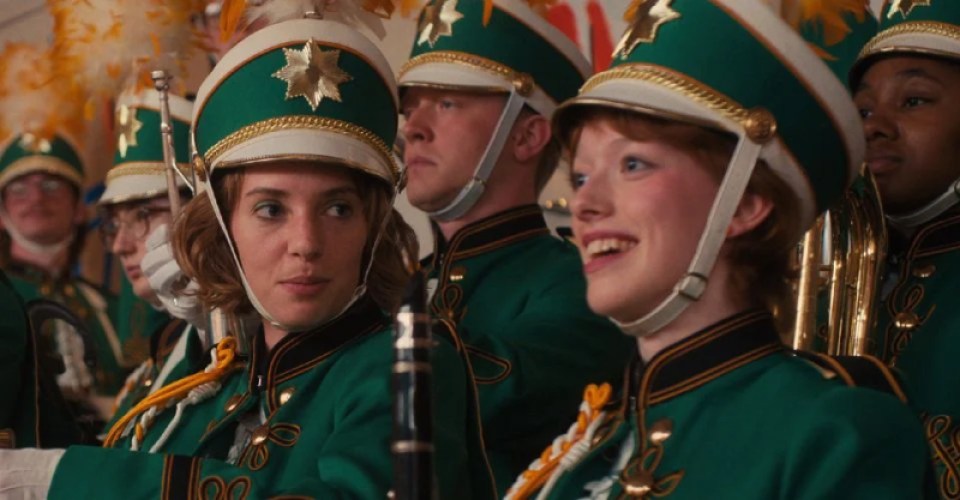 In a still from Stranger Things, Robin and Vickie are sitting side by side in their 1980s green marching band uniforms. Vickie is smiling off to the left of cameras and Robin is staring upon the side profile of Vickie's face intensely.