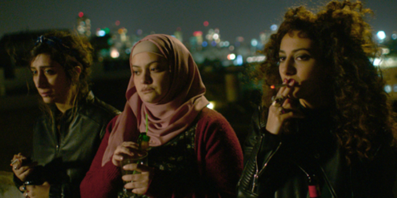 Three women stand on a roof drinking, two smoke cigarettes, the third is wearing a hijab