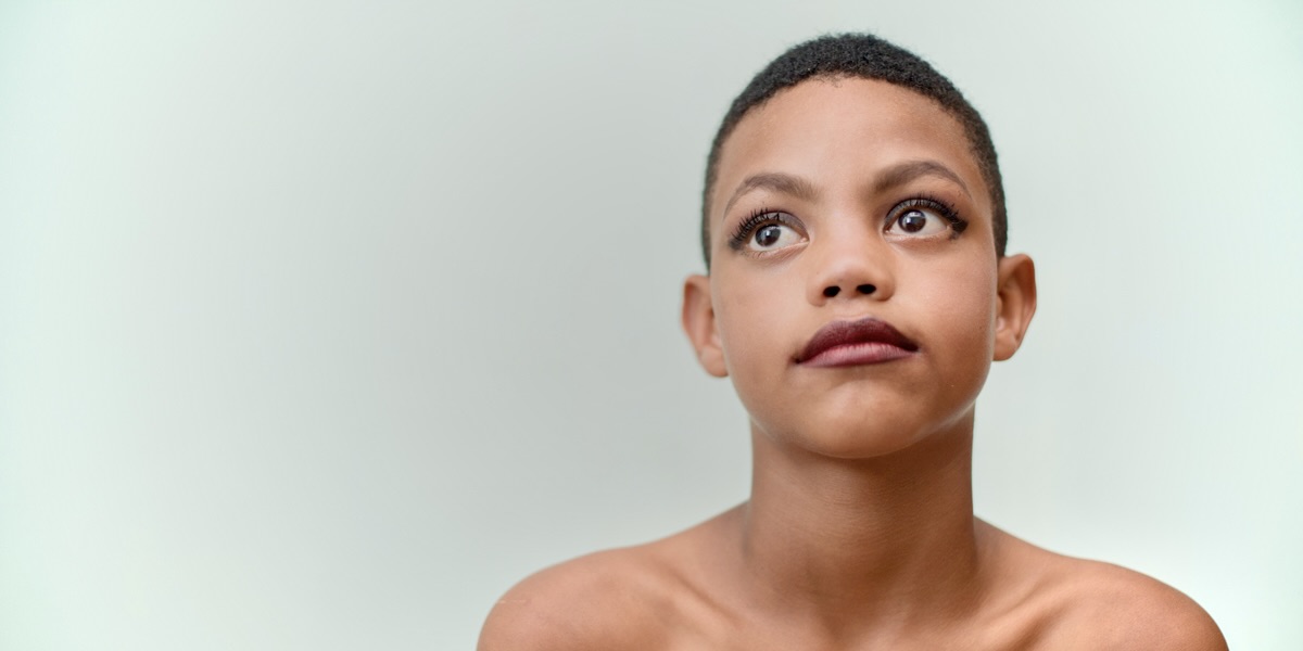 A young Black trans child with short hair and dark purple lipstick looks up and to the left of camera, in front of a smoky grey background