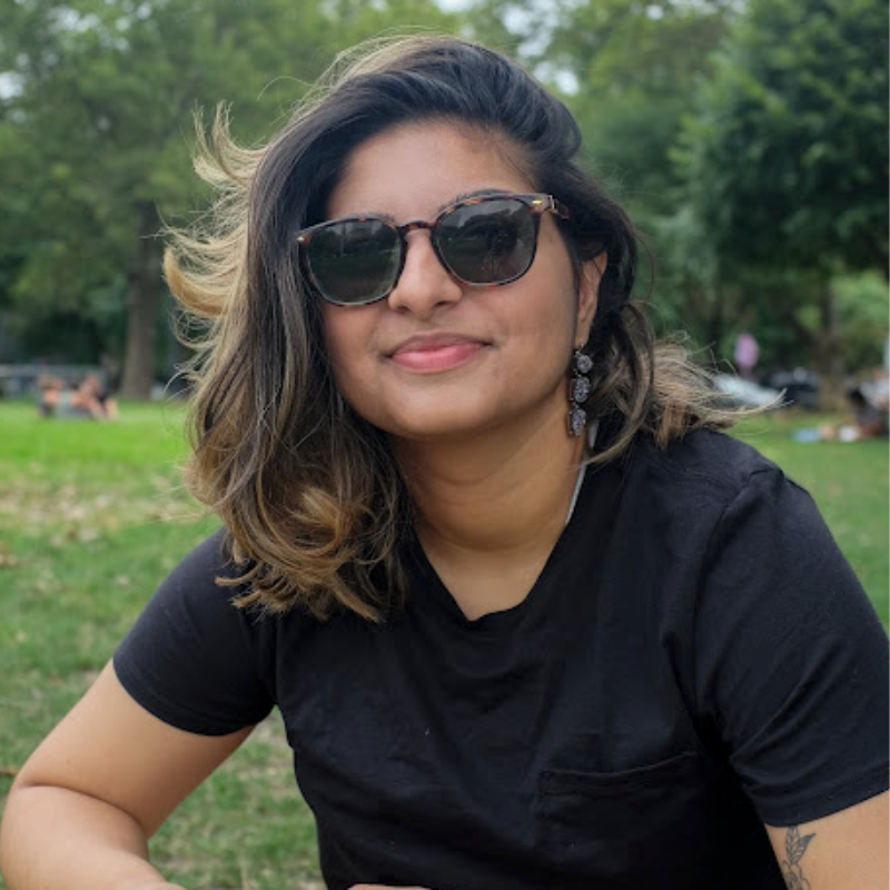 Ashni is a South Asian person with shoulder length hair in black sunglasses and black shirt sitting outside on the grass