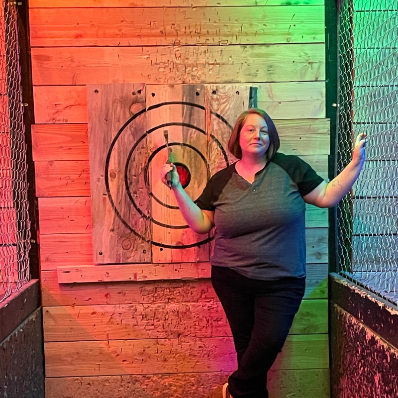 Darcy is a white person with a brunette bob in a grey and black henley shirt, standing in front of a dart board that is lit in rainbows.