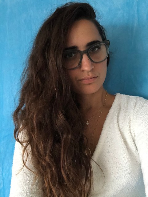 Kayla Kumari Upadhyaya is a brown dyke with long brown curly hair and a white fuzzy shirt on. She wears blue glasses. She probably has "lesbian eyebrows."