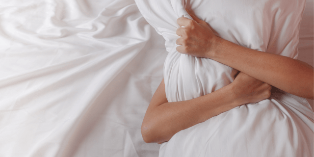 A person with light skin lying on white sheets, hugging a white pillow, which is obscuring their face