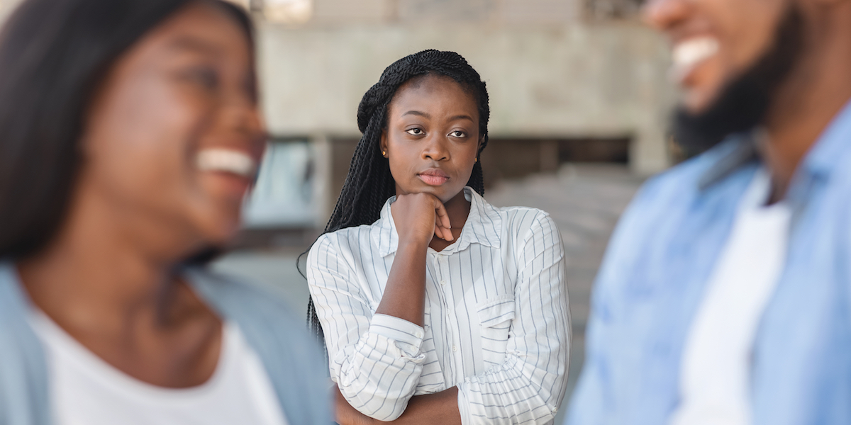 a Black woman with long braids stands in the middle of the frame, looking upset, while watching a Black man and woman laugh together in the front of the frame