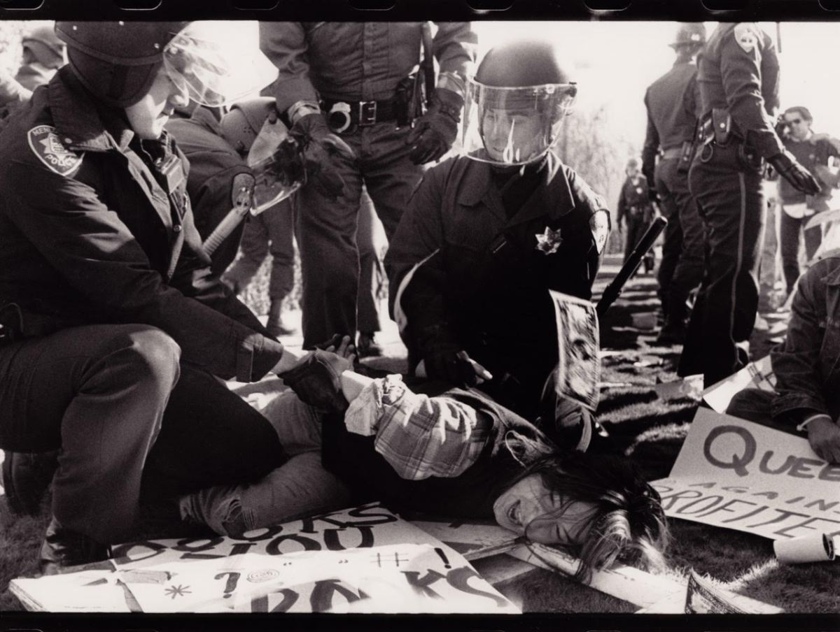 A black and white photo shows a woman with long, dark hair wearing a flannel and sweater vest on the grass on her side. Her hands are being cuffed behind her back by cops in riot gear. Protest signs litter the ground.