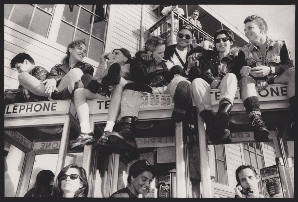 A black and white photo shows seven smiling queer women sitting on top of telephone booths. Three people stand below them. Two others are visible on a balcony behind them.