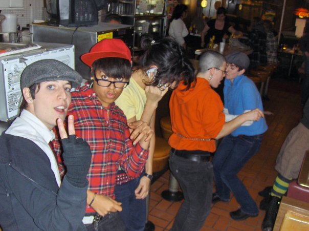 From left to right: Kate, a white person with black hair, wears a button up shirt, a red tie, a newsboy cap and black fingerless gloves. They're making a peace sign. Khaleb, a Black person with short, straight black hair that covers their forehead, wears a red flannel, a red cap and glasses while dancing at the camera. Pidgeon, who has light brown skin and long black hair, wears a yellow shirt and talks on a small, silver phone; their face is not visible. Peter, a white person with a shaved head wearing a red, button-up shirt and black pants, dances with Mica, a white person with short, curly brown hair, who's wearing a newsboy cap, a blue polo shirt and black jeans. Standee's diner and some other diner guests are visible in the background.