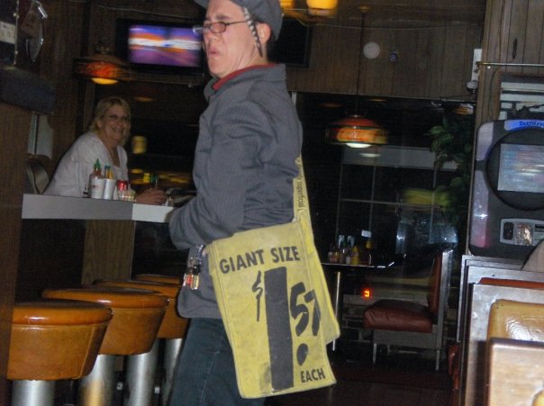 Peter Wilde, a white person wearing a grey jacket and a grey newsboy cap with a feather on it, scrunches up their face while they dance and lip sync in Standee's diner. They have burlap bag over their shoulder that reads, "Giant Size - $1.57 each." The counter at Standee's Diner is visible in the background. Tinkerbell, a middle-aged server with shoulder-length blonde hair who is wearing a white shirt, leans over the counter and smiles at Peter. 