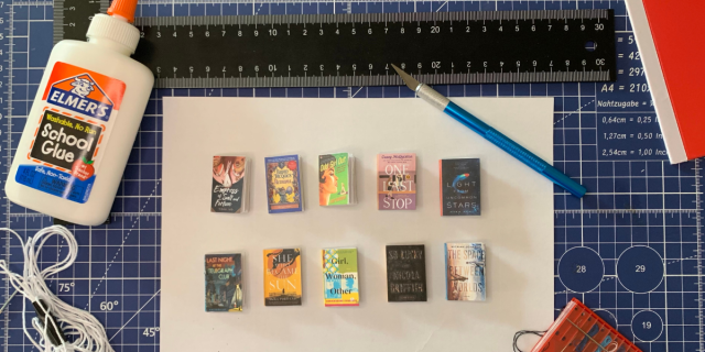 A series of tiny replica novels laid out on a bright blue cutting mat with craft supplies splayed out around them