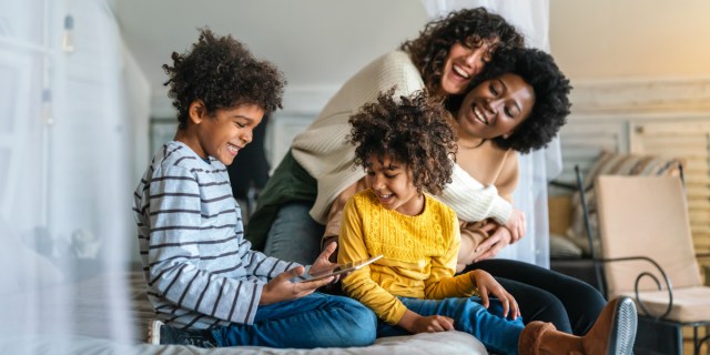 Lesbian moms — one a woman with curly brown hair and one a Black woman with an afro — smile while their two children, both mixed race boys with curly hair, play on their tablet. Their home is well lit with white walls.