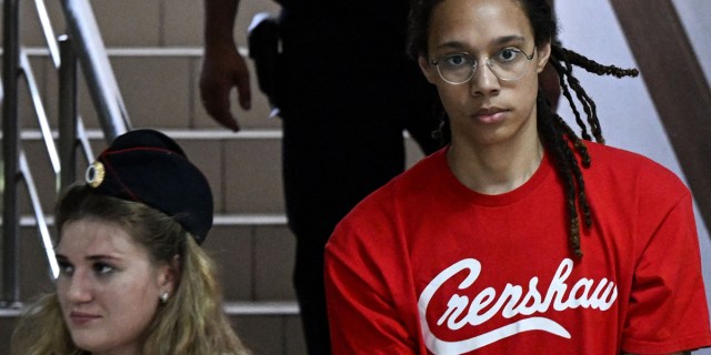 Brittney Griner is in a red t-shirt that says "Crenshaw" and round glasses, descending on a staircase in a government building. In front of her is a w white woman with blonde hair, wearing a hat that looks like that of an Russian official or security officer.