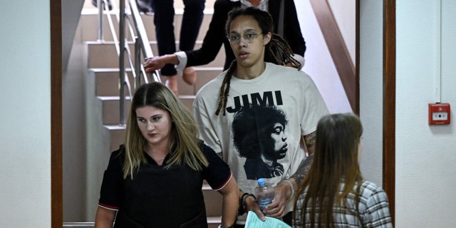 Brittney Griner is in a Jimi Hendrix shirt, walking down stairs, in a plain government building. In front of her is a white woman with blond hair and a black polo t-shirt.