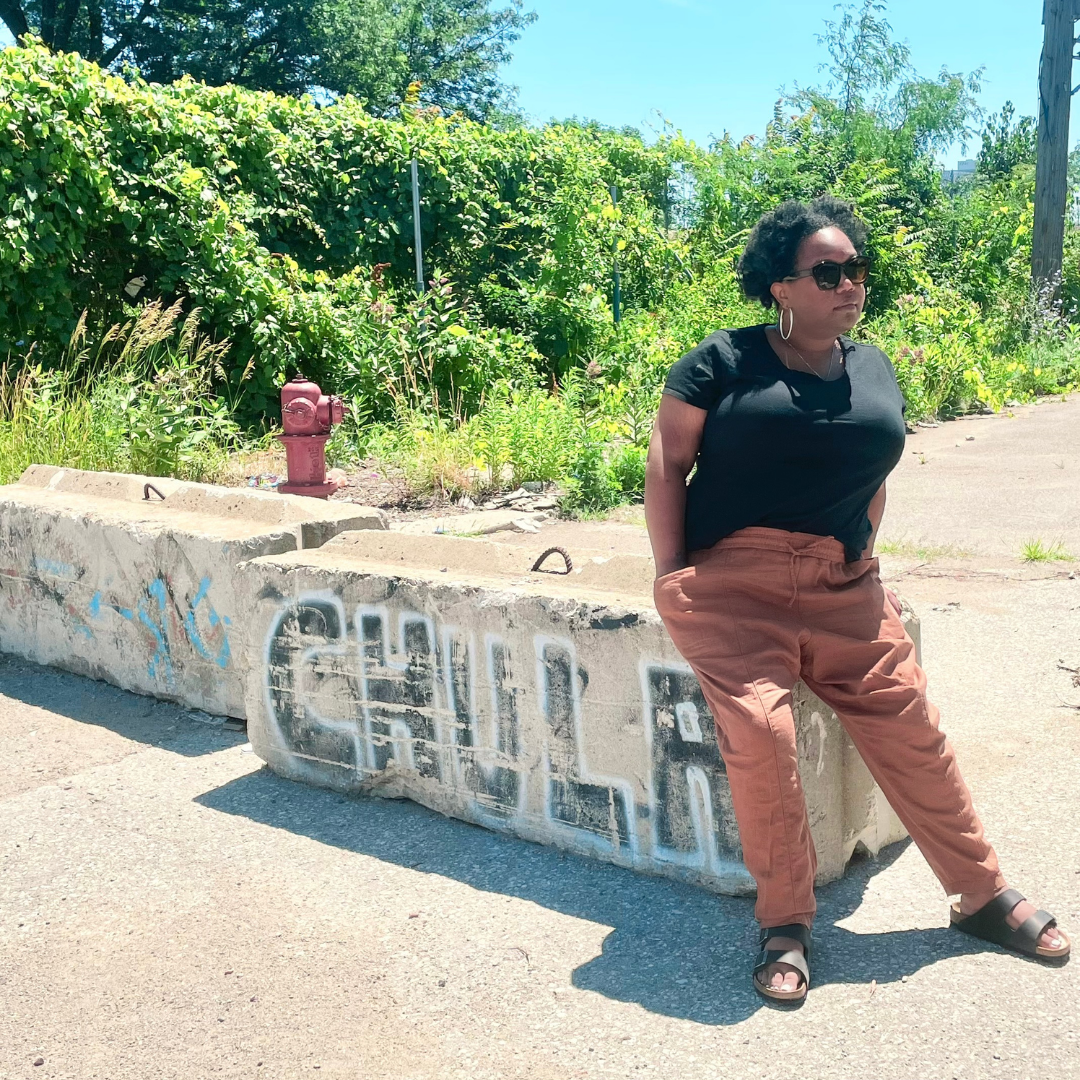 Carmen, a Black woman, sits against a concrete block graffitied with the word, "chula." She is wearing coral pants, birkenstocks and a black tee. Her hair is parted and curly and she is wearing hoop earrings and sunglasses.