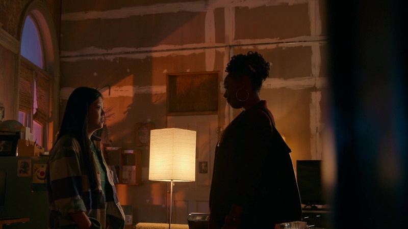 Malika and Alice, standing facing each other in Alice's loft. Alice is wearing a long sleeve flannel shirt and Malika is wearing a blazer. A lamp is placed between them.