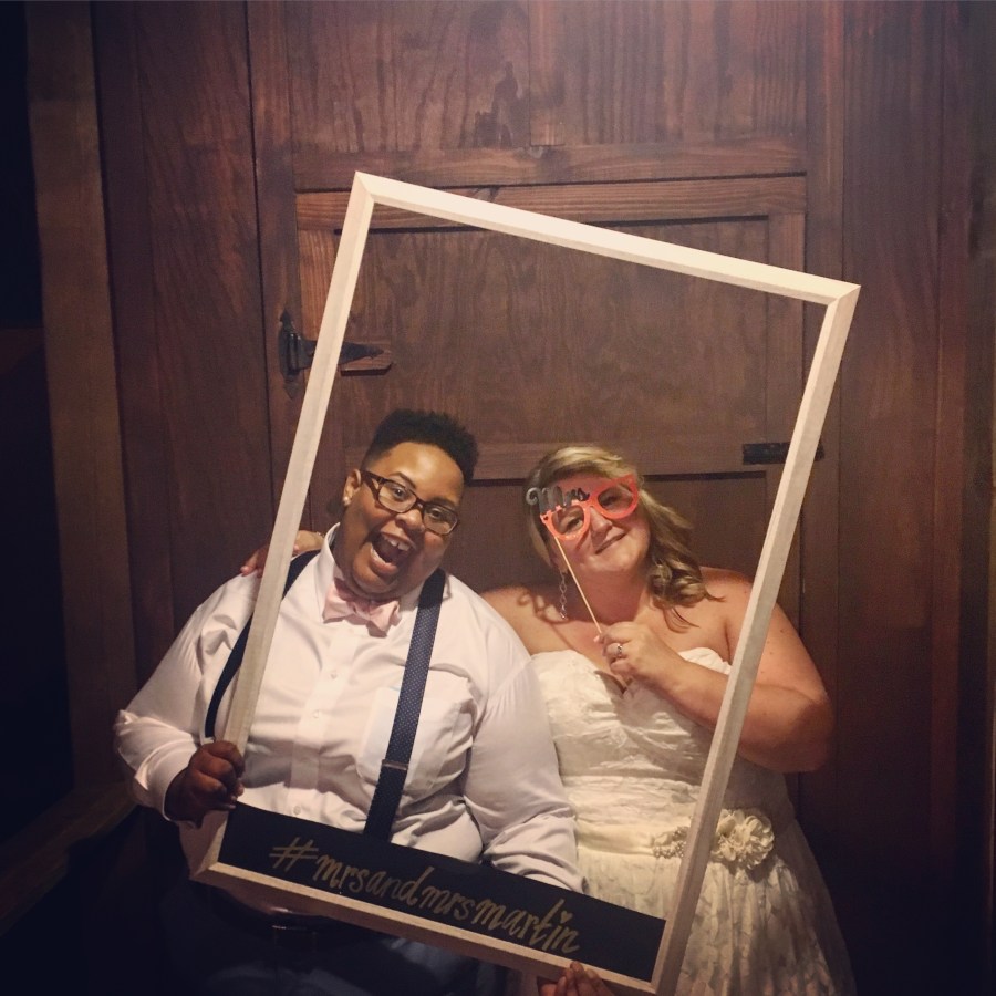 a wedding photo of shea and Jane. shea is wearing a white shirt, pink bowtie, suspenders, glasses, navy pants and a huge smile. Jane is wearing a gorgeous lacy wedding dress and is holding up glasses that are the type for photo props. shea is holding up a polaroid esque wooden frame with #mrsandmrsmartin written on its base. shea is a Black nonbinary human with short curly black hair and Jane is a white woman with shoulder length blonde hair that is curled and falling down to her shoulders
