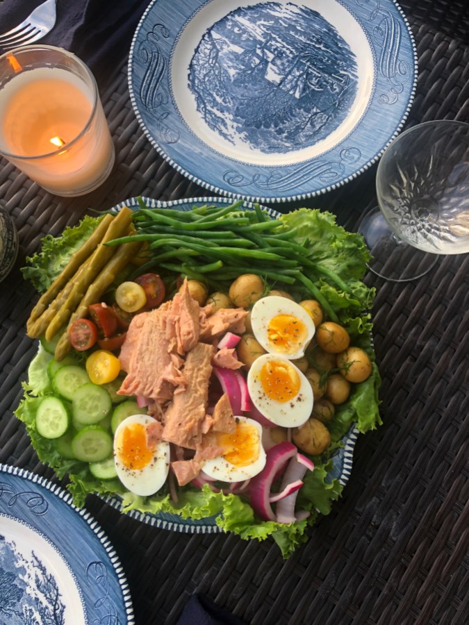 A tuna nicoise on a blue and white plate. The salad features green beans, pickled asparagus, medium boiled eggs, pickled red onions, baby potatoes with dill, sliced cucumber, sliced multicolor cherry tomatoes, green beans, romaine lettuce, and a pile of tuna. There is a candle lit on the table and a coupe glass of wine.