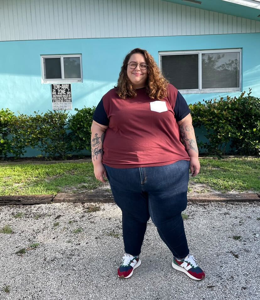 a photo of stef rubino. stef is white human with medium long brown wavy hair wearing a color block tee and jeans, standing in front of a blue building. stef has glasses.
