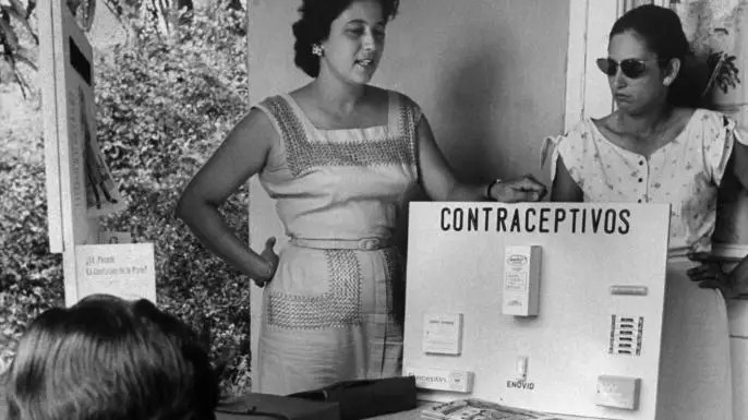 A black and white image of two Puerto Rican women in 1960 standing next to a poster board that reads "Contraceptivos" (contraceptives) while giving a presentation.