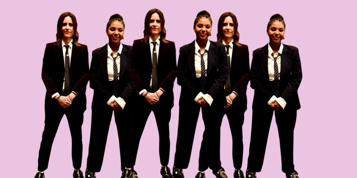 Kate Moennig and Jordan Hull stand in suits with their hands clasped in front of them against a pink background.